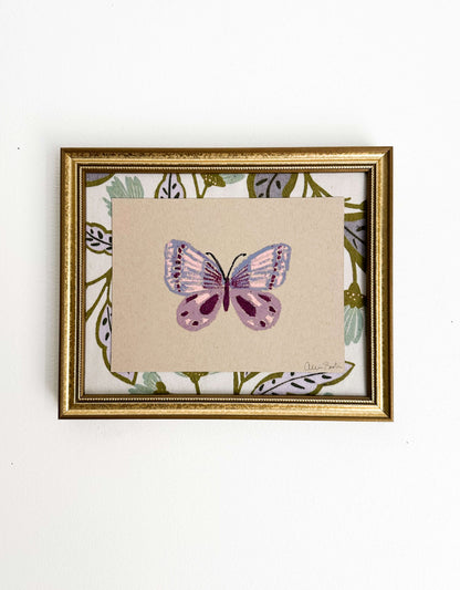 A painting of a purple butterfly on tan paper and mounted on floral fabric. The painting is in a gold frame and shown on a white wall.
