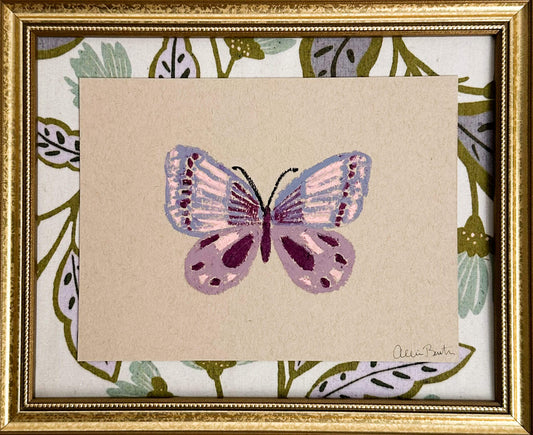 A painting of a purple butterfly on tan paper and mounted on floral fabric. The painting is in a gold frame.