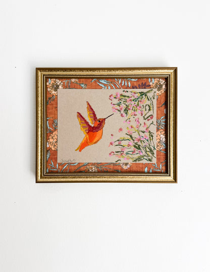 A painting of an orange hummingbird and pink flowers on tan paper. The painting is in a gold frame on a white wall.