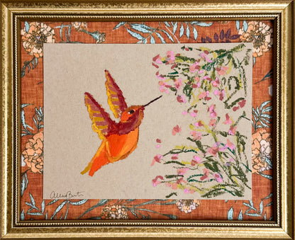 A painting of an orange hummingbird and pink flowers on tan paper. The painting is in a gold frame.
