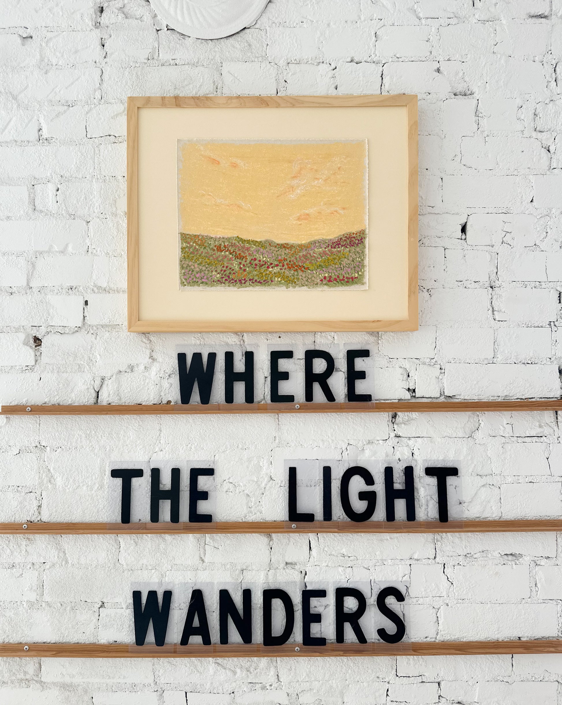 Framed artwork on a white brick wall with 'WHERE THE LIGHT WANDERS' text below.