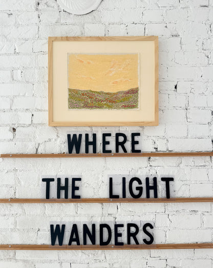 Framed artwork on a white brick wall with 'WHERE THE LIGHT WANDERS' text below.