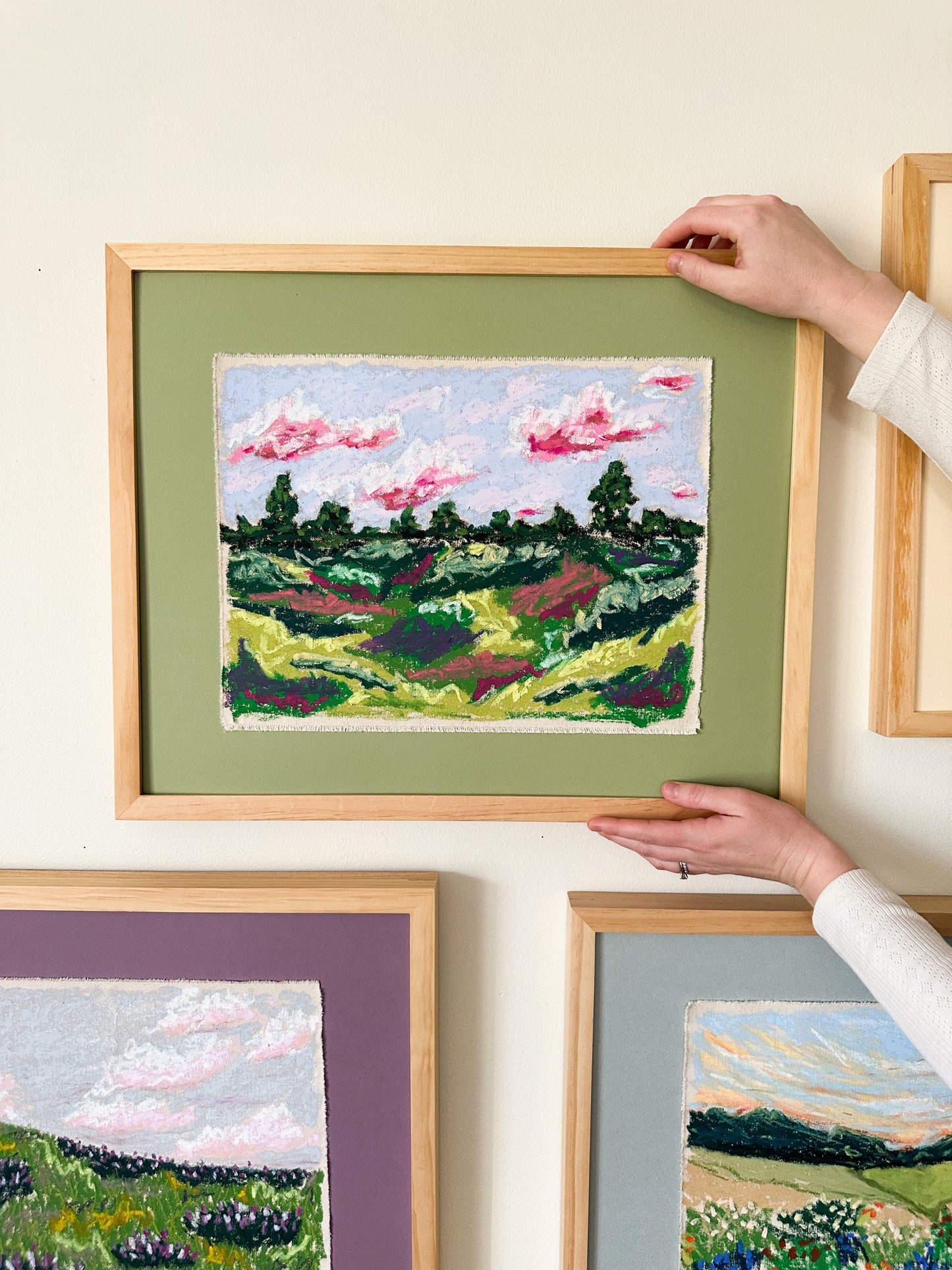 Person hanging a framed landscape painting on a white wall with other landscape paintings.