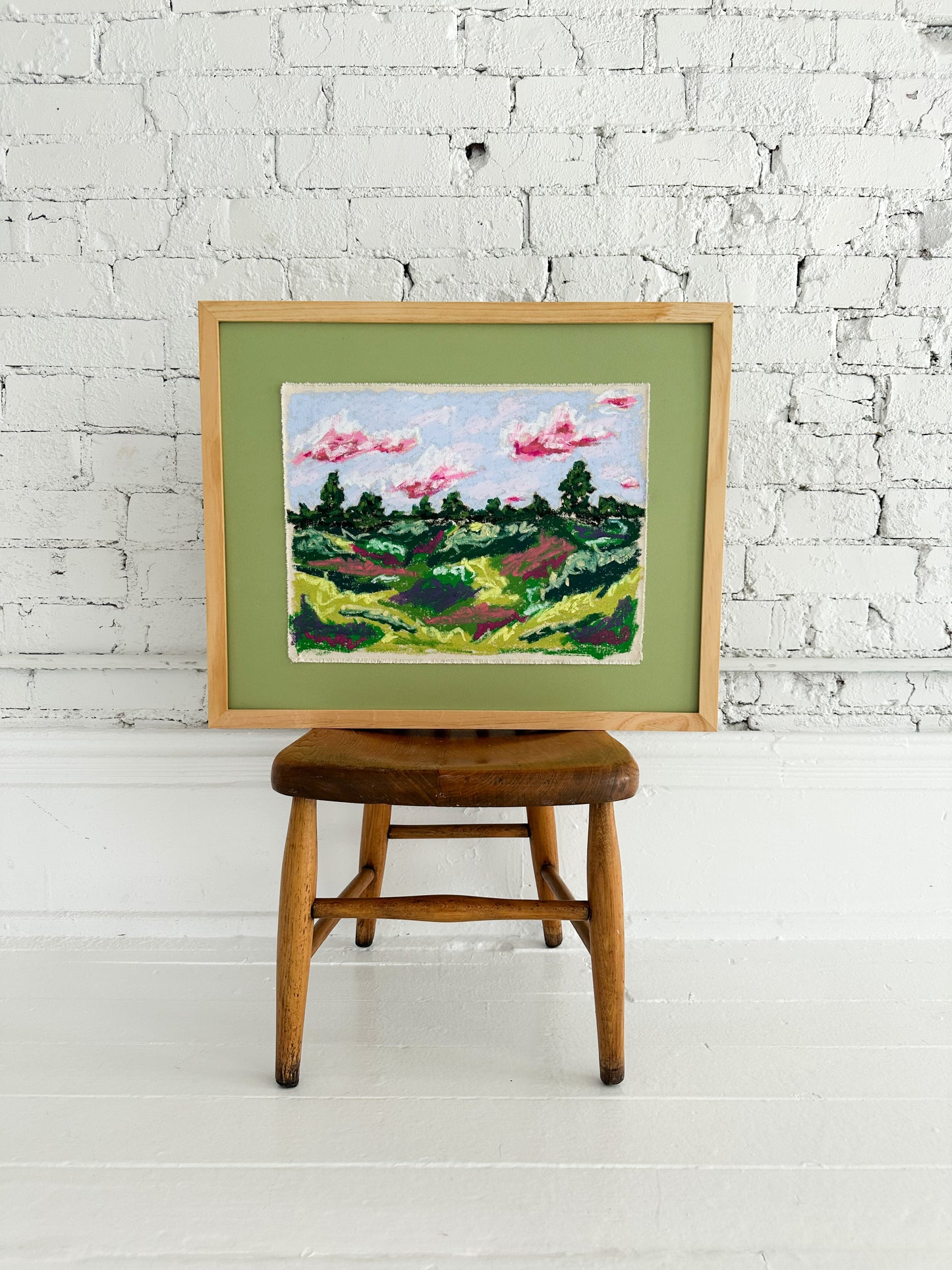 Framed painting of a landscape on a wooden stool against a white brick wall.