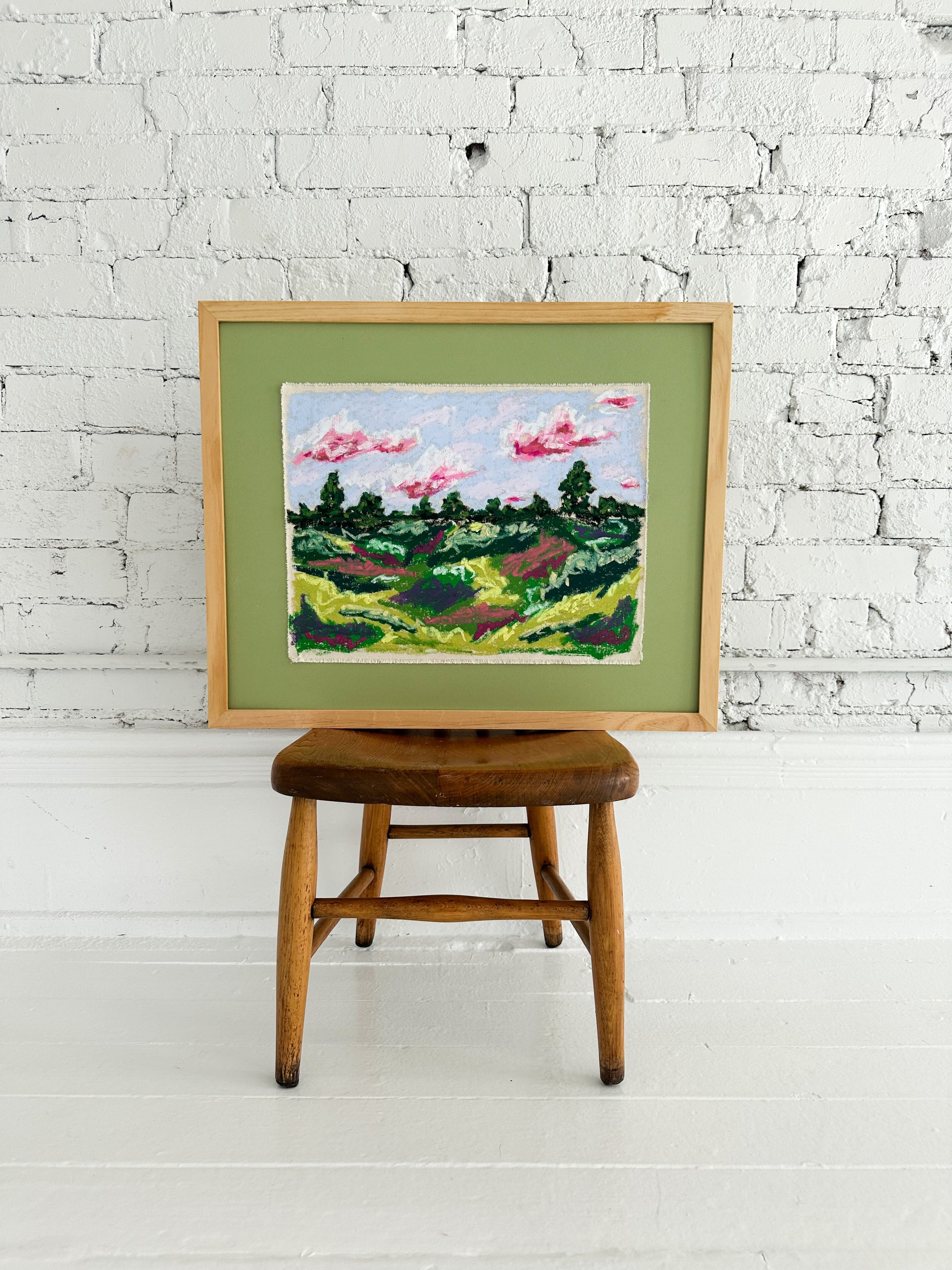 Framed painting of a landscape on a wooden stool against a white brick wall.