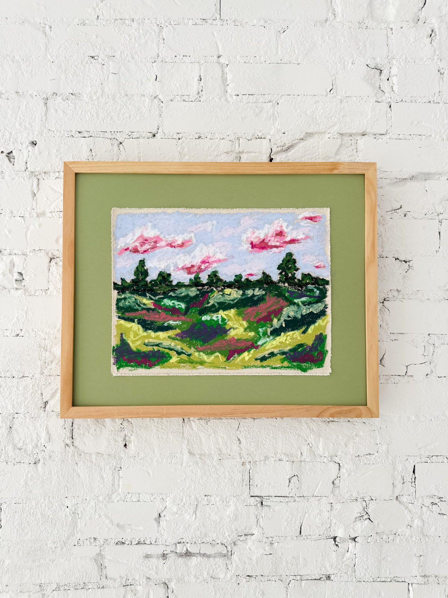 Framed artwork of a landscape with trees and pink clouds on a white brick wall.