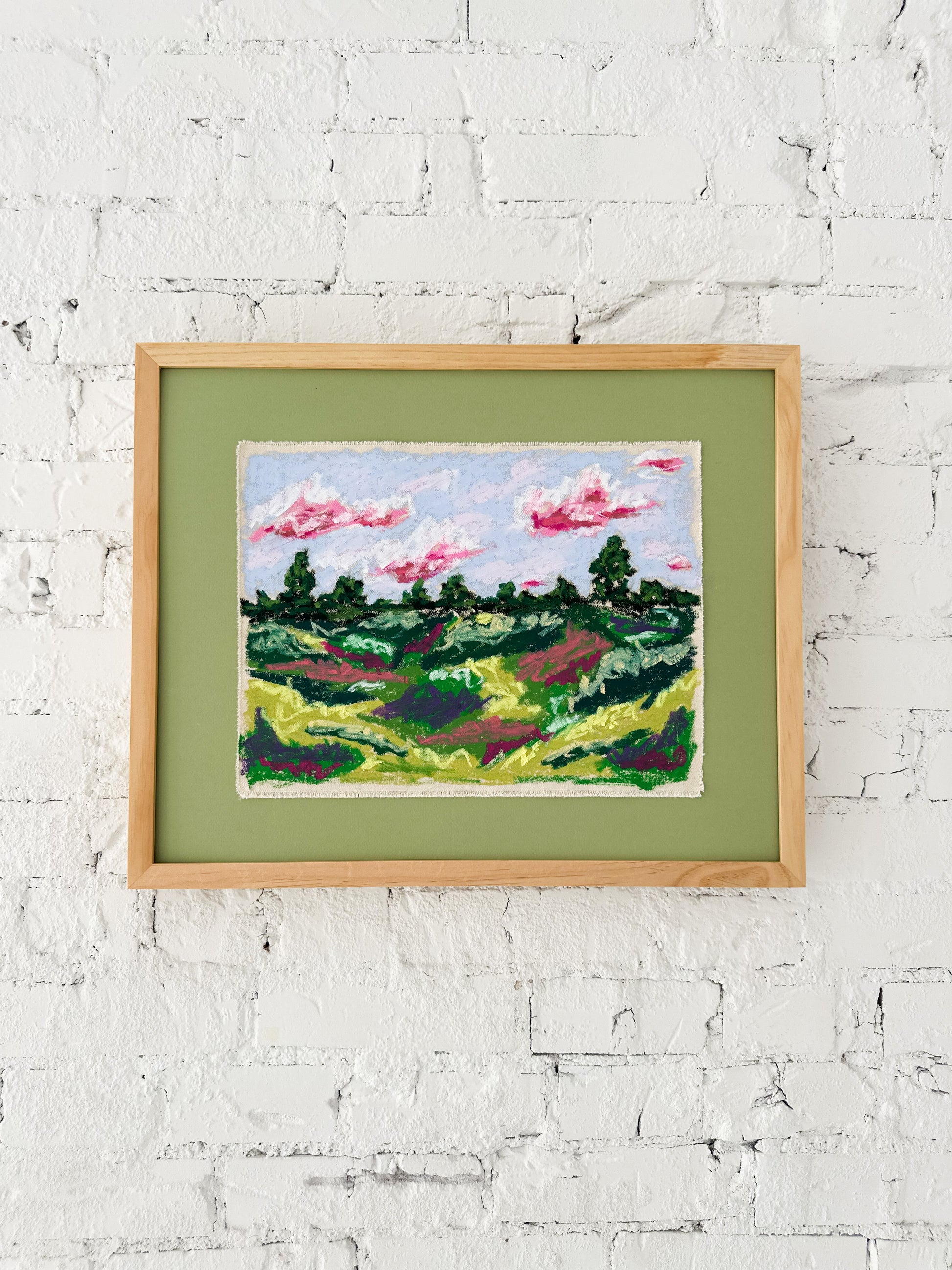Framed artwork of a landscape with trees and pink clouds on a white brick wall.