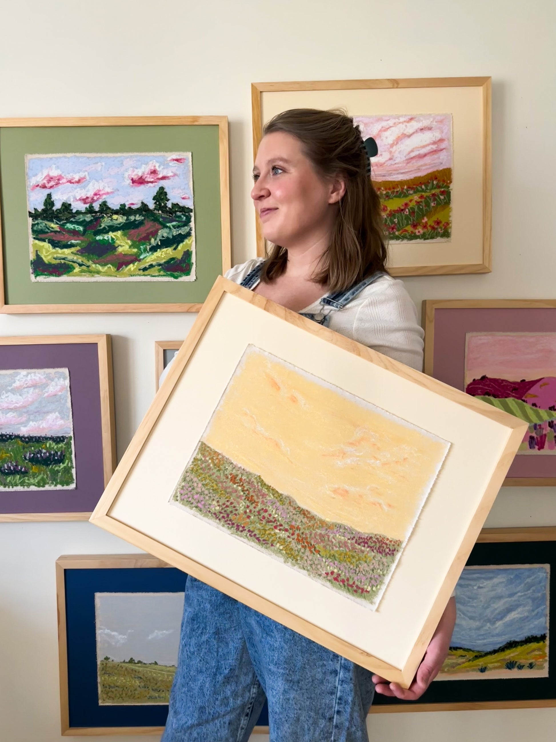 Woman holding a framed landscape painting in front of other framed artworks on a wall.