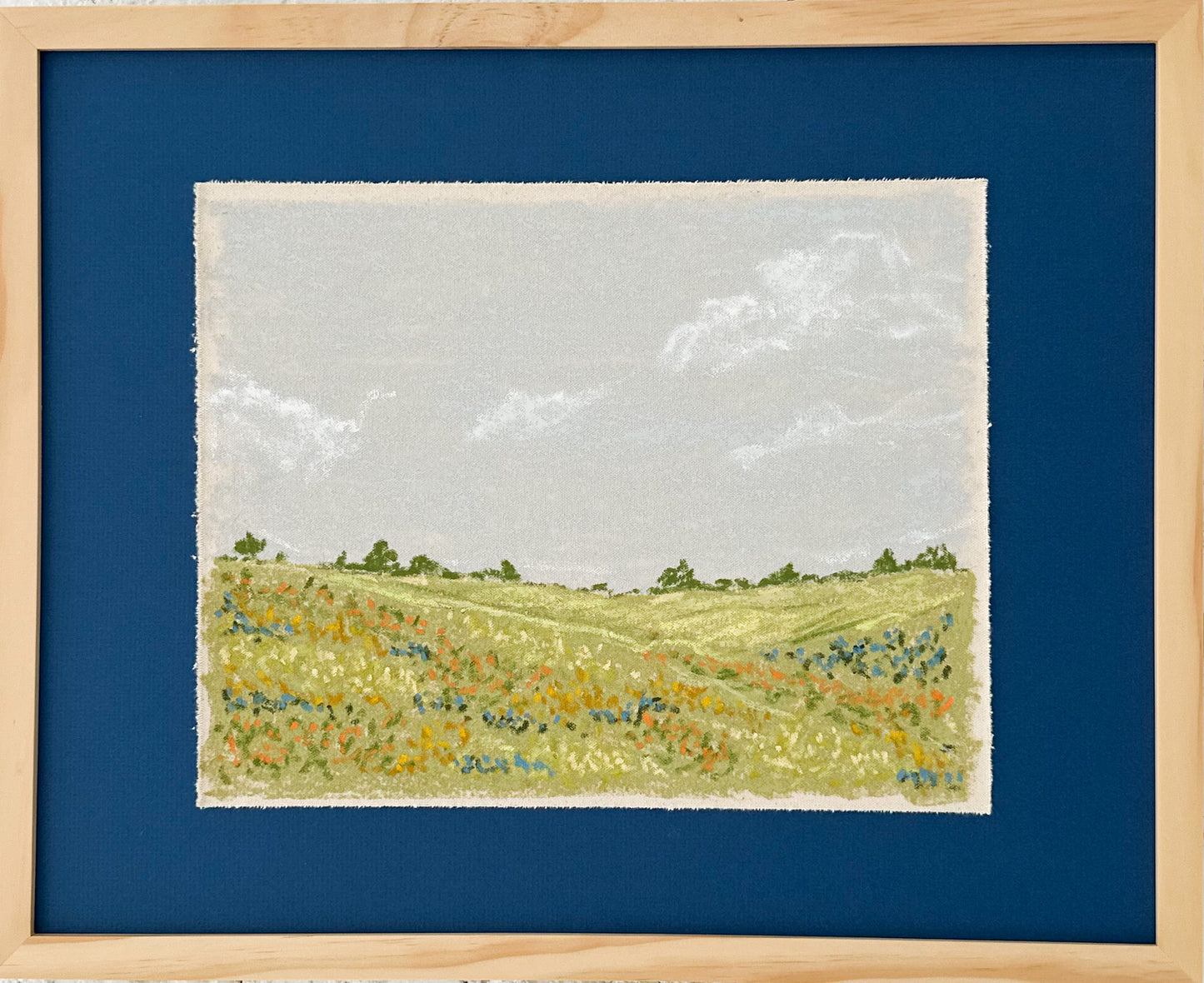 An oil pastel painting depicting green hills with orange, blue and yellow wildflowers. There are trees in the background with a dusty blue, cloudy sky.