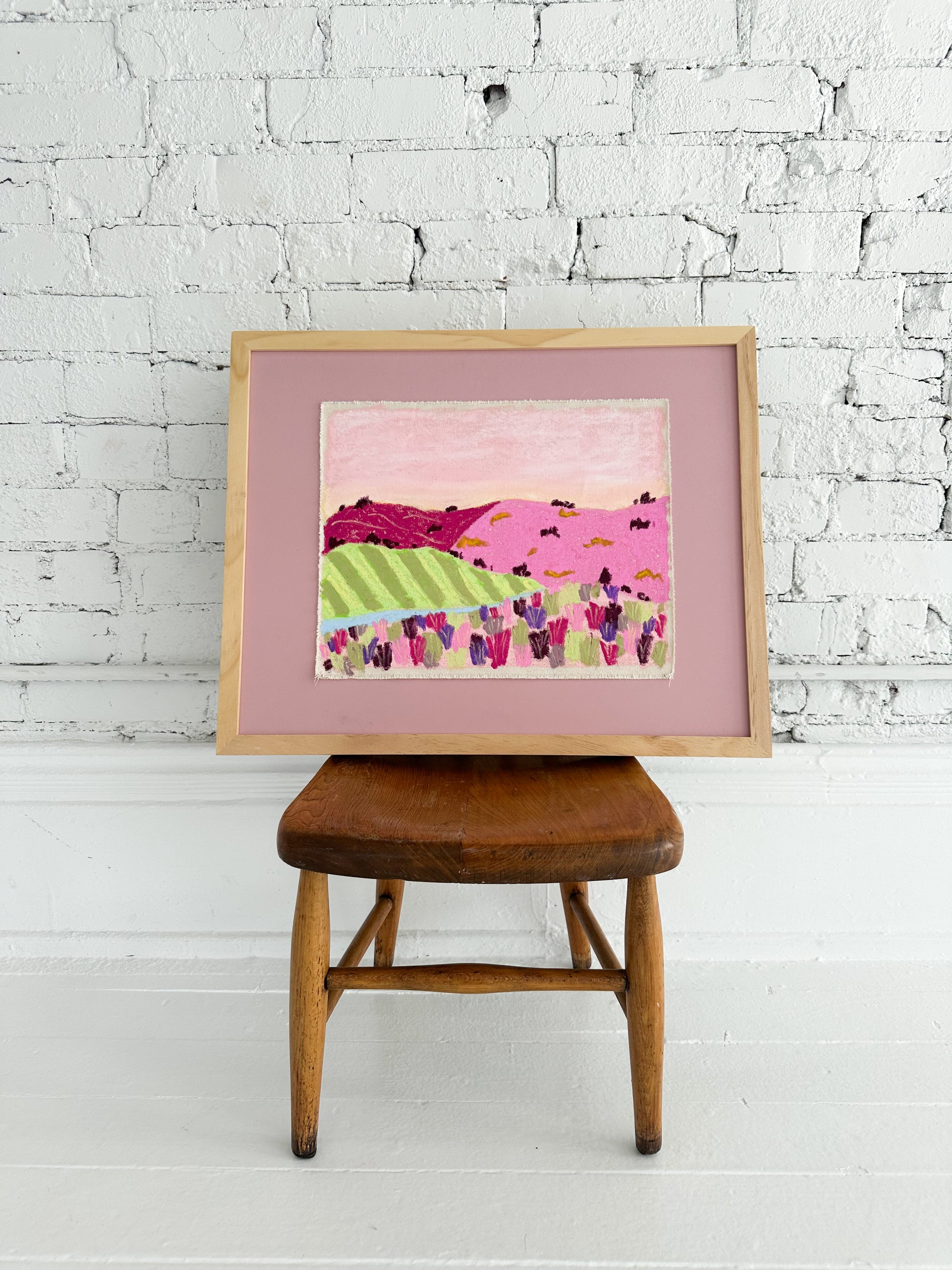 Framed colorful abstract painting on a wooden stool against a white brick wall.