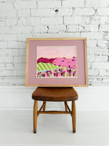 Framed colorful abstract painting on a wooden stool against a white brick wall.