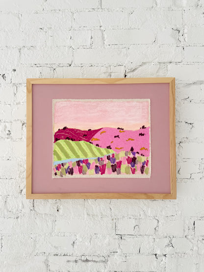 Framed landscape artwork with pink and green hills on a white brick wall.
