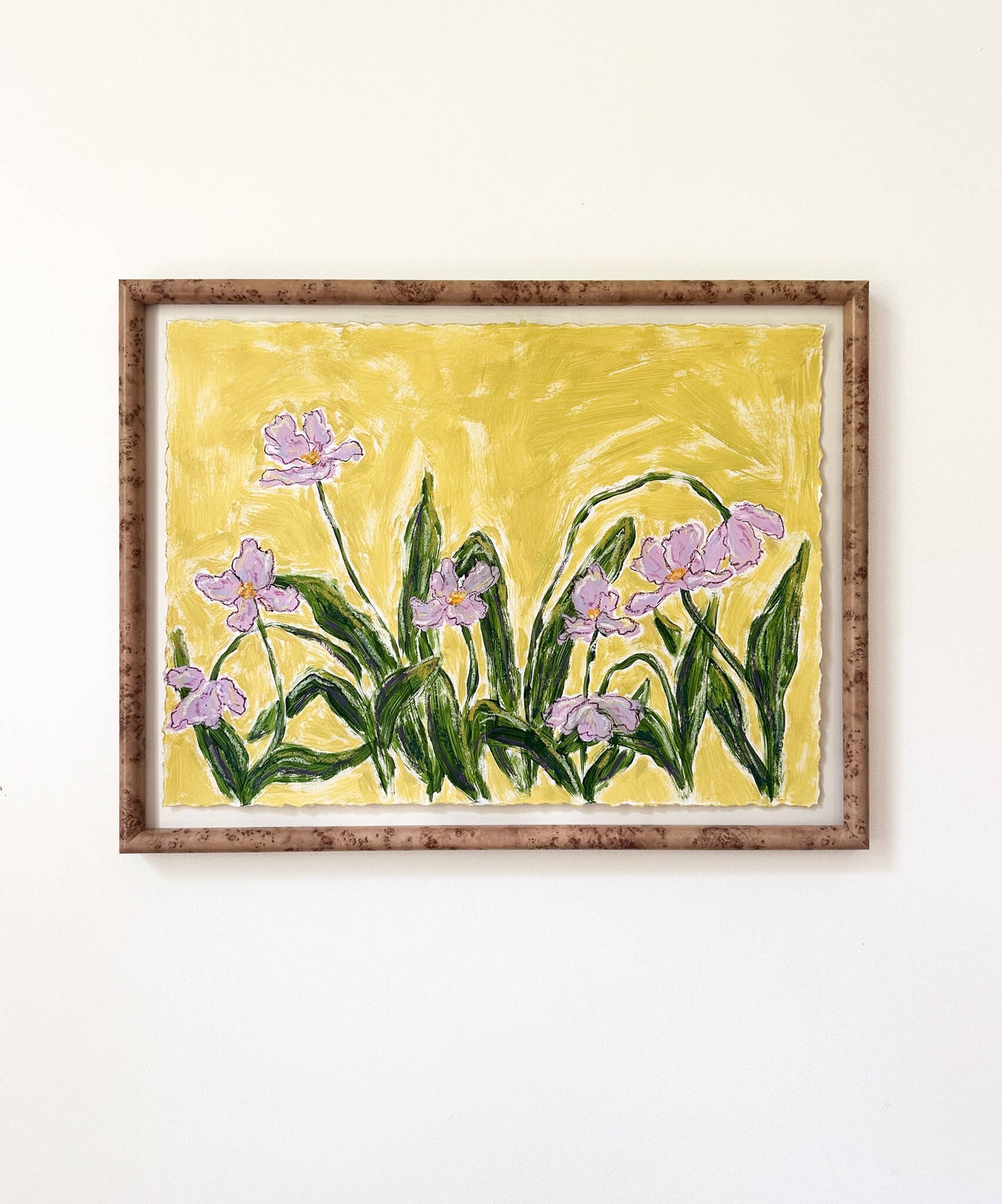 a framed painting of light purple flowers with green stems painted on a bright yellowish green background and hung on a white wall.