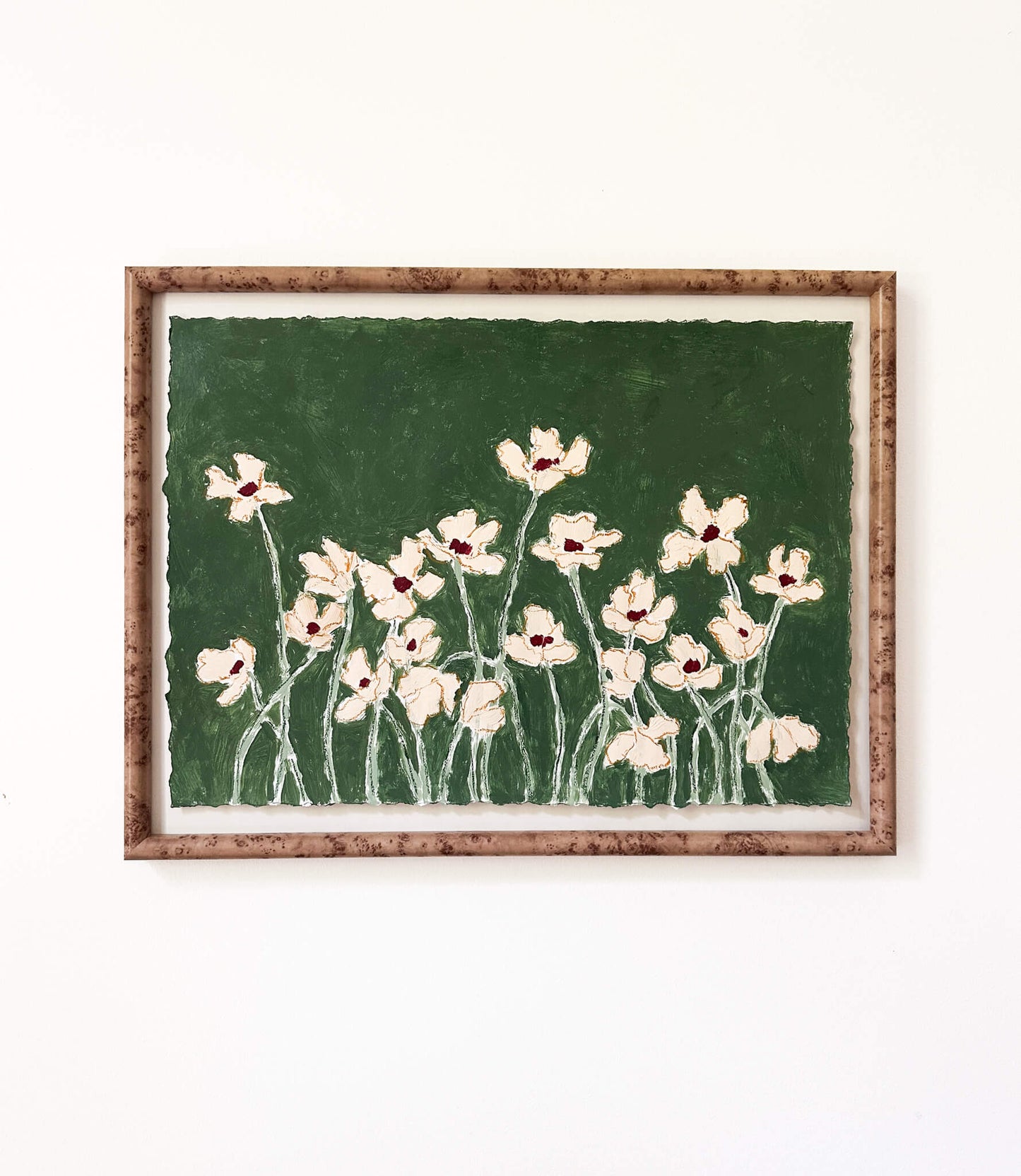 a framed painting of cream flowers with green stems painted on a dark green background and hung on a white wall.