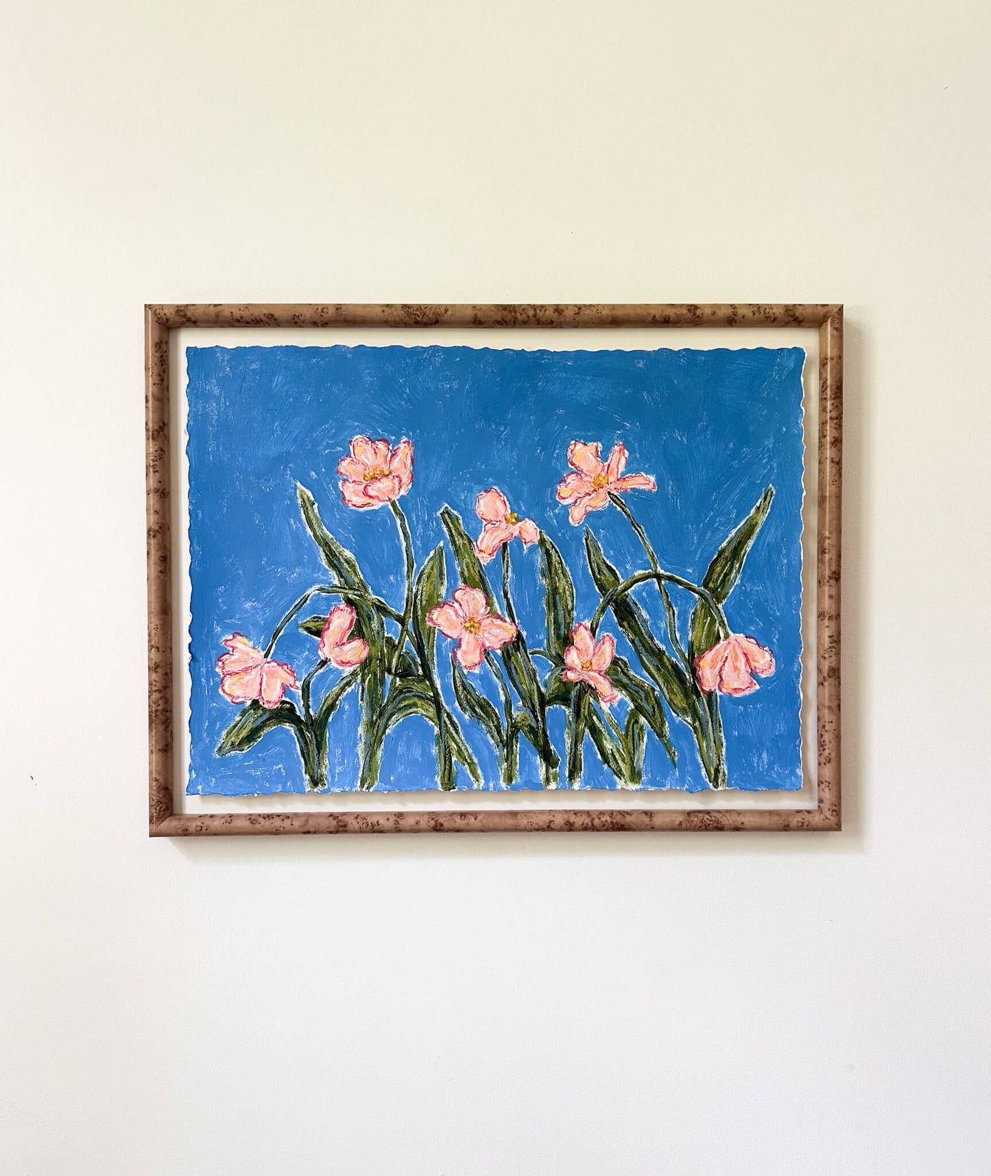 a framed painting of pink flowers with green stems painted on a bright blue background and hung on a white wall.