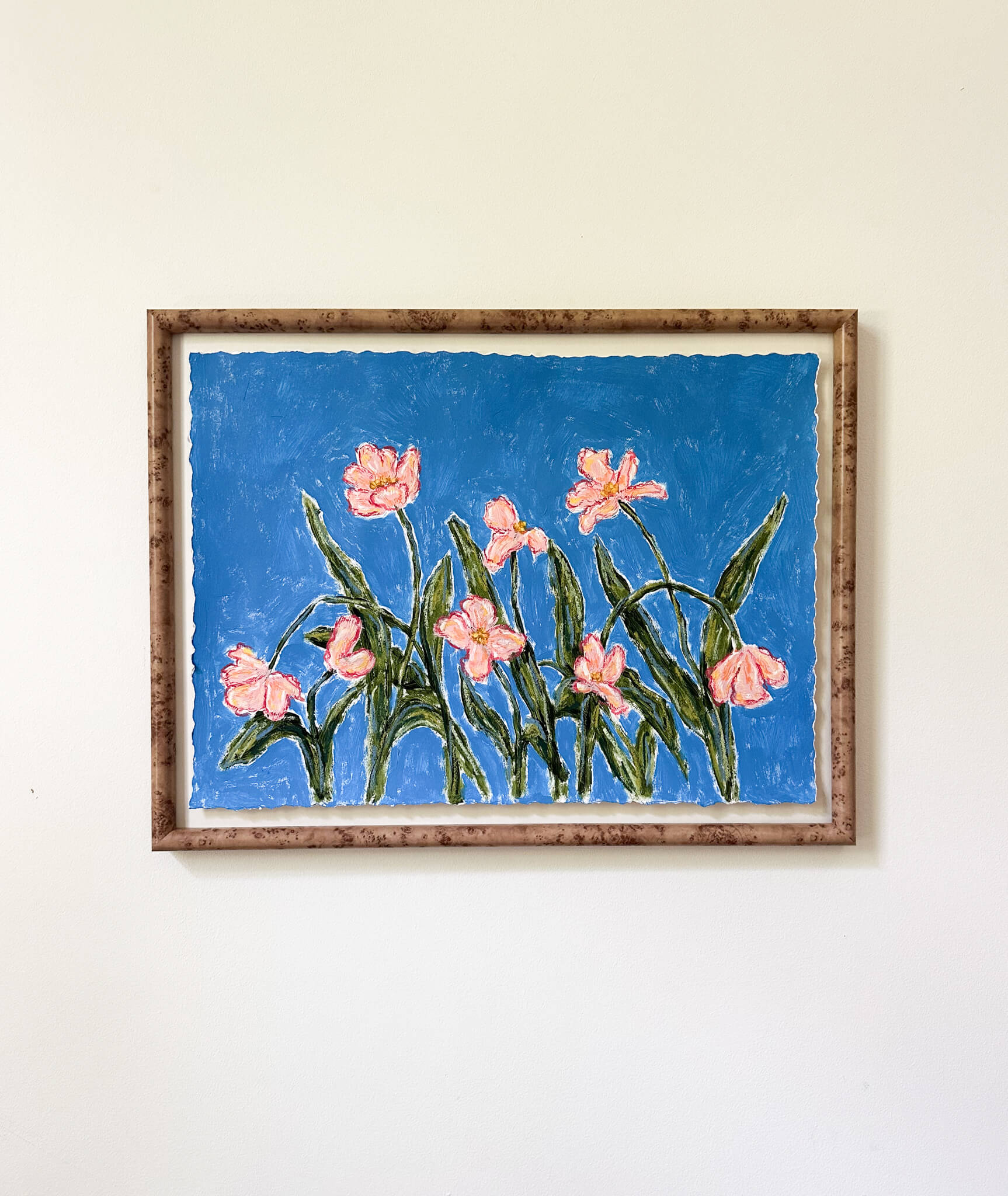 a framed painting of pink flowers with green stems painted on a bright blue background and hung on a white wall.