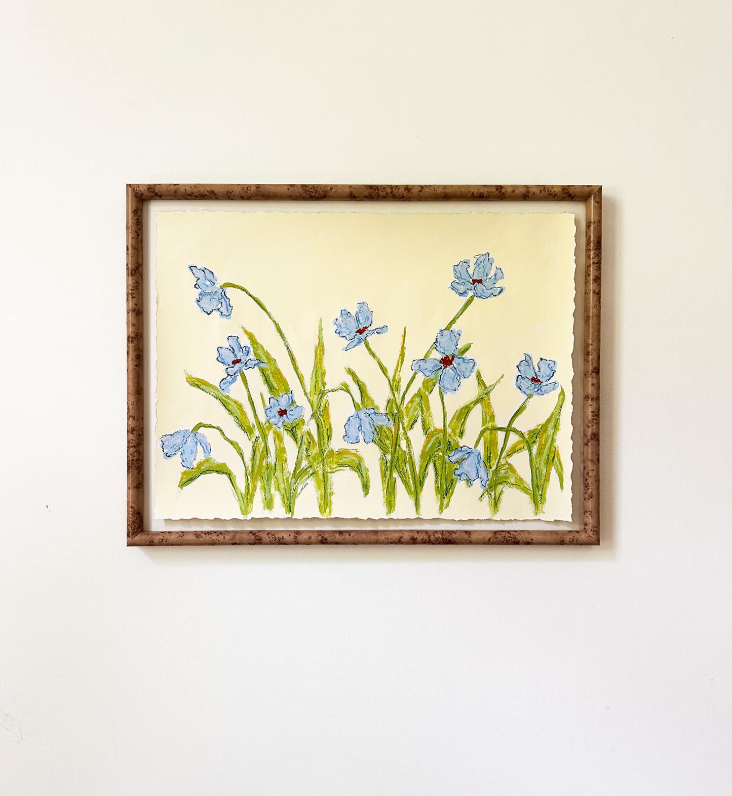 a framed painting of light blue flowers with green stems painted on a light yellow background and hung on a white wall.
