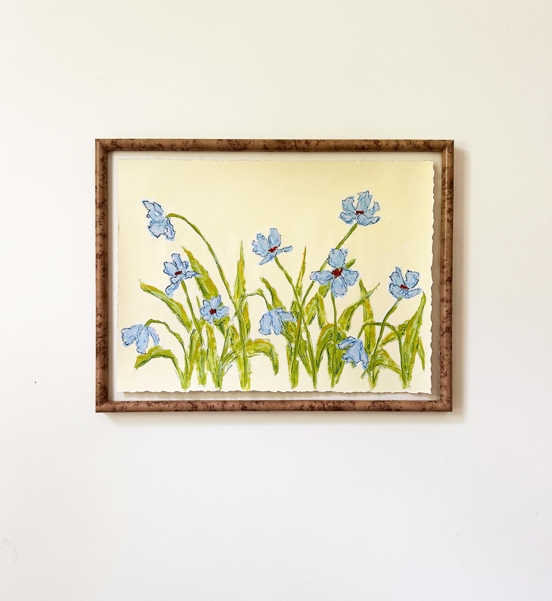 a framed painting of light blue flowers with green stems painted on a light yellow background and hung on a white wall.