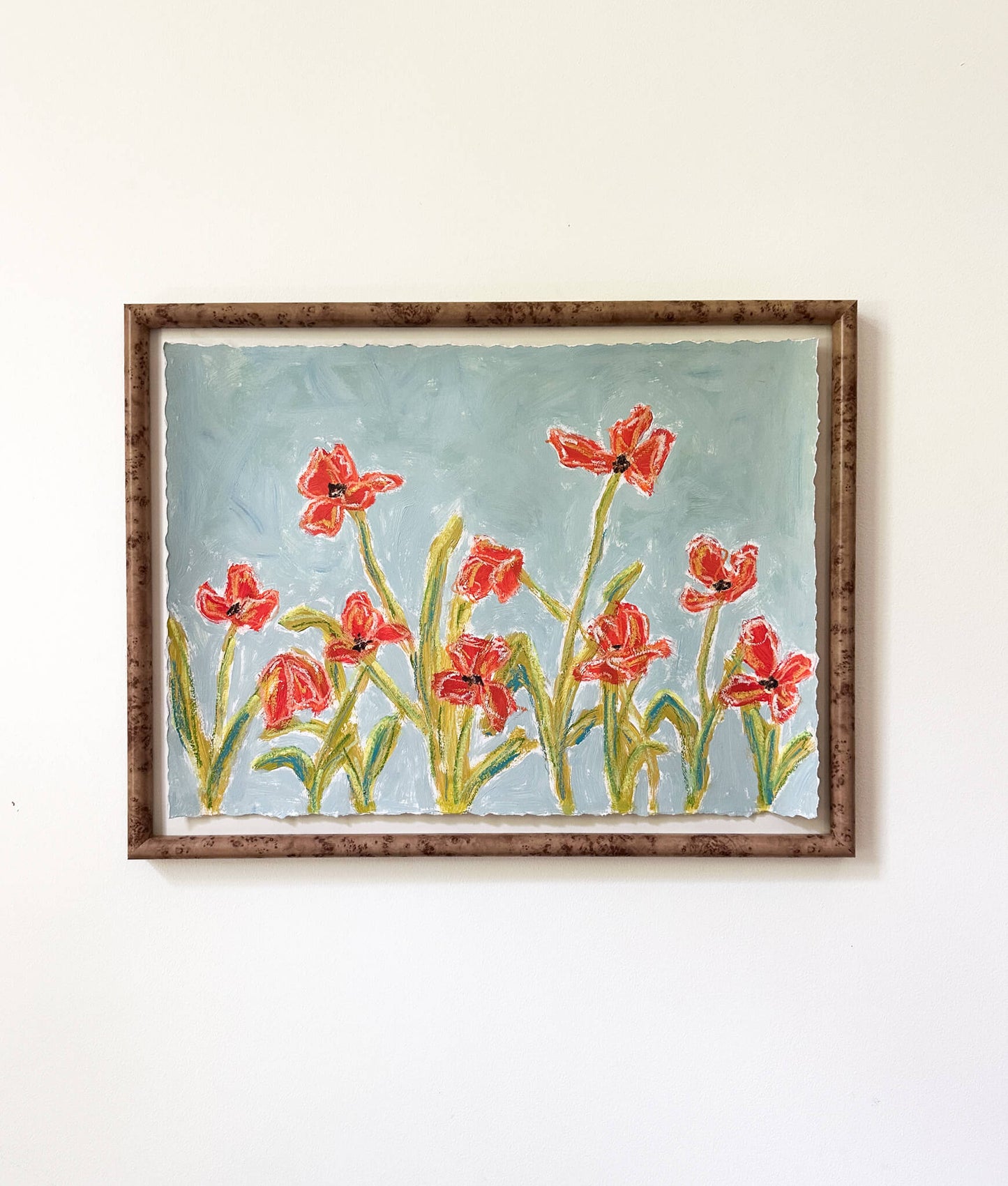 a framed painting of orange flowers with green stems painted on a dusty blue background and hung on a white wall.