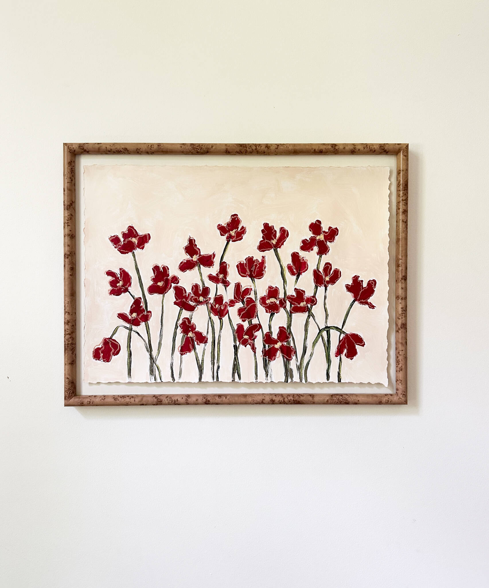 a framed painting of dark red flowers with green stems painted on a beige background and hung on a white wall.