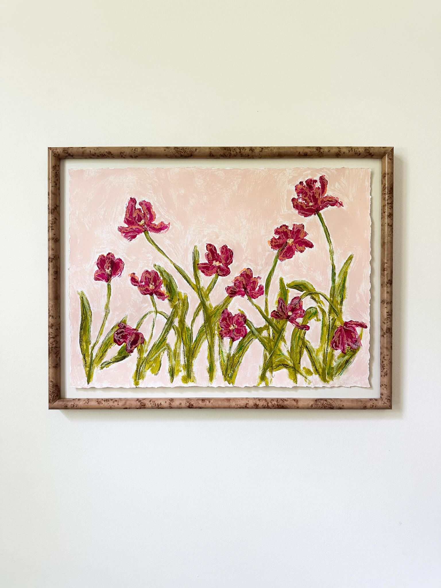 a framed painting of dark pink flowers with green stems painted on a light pink background and hung on a white wall.
